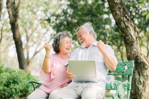 A senior adult couple on a park bench, celebrating while holding a tablet