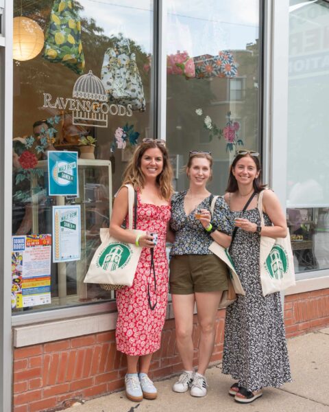 Three people smiling outside of Ravensgoods, holding Malt Row on Damen tote bags