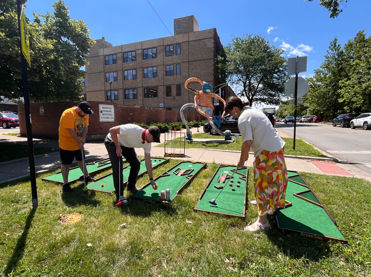 Three people playing mini golf on PutterBird made putting greens, situated in front of the sculpture Blind Love.
